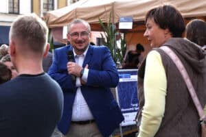 Mężczyzna na tle stoisk wystawienniczych pozuje do fotografii uśmiechając się do aparatu. W lewej dłoni trzyma mikrofon