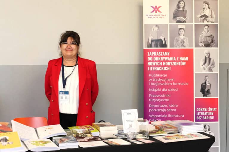 Kobieta w czerwonej marynarce i okularach stoi za stołem pełnym książek i materiałów promocyjnych. Po jej prawej stronie znajduje się wysoki baner reklamowy z logo "Wydawnictwo Trzecie Oko" i hasłem "ZAPRASZAMY DO ODKRYWANIA Z NAMI NOWYCH HORYZONTÓW LITERACKICH!" oraz informacją o publikacjach w tradycyjnym i brajlowskim formacie