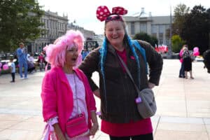 Plac Litewski w Lublinie. Dwie szeroko uśmiechnięte kobiety stojące na placu. Kobieta po lewej ma różową perukę, różowy kapelusz z falbanami oraz różową kurtkę i torebkę. Kobieta po prawej ma dwa niebieskie warkocze i dużą różową opaskę w białe kropki przypominającą uszy Myszki Miki, a także ciemną kurtkę i różową koszulkę. W tle widać innych uczestników zgromadzenia i historyczne budynki miasta.
