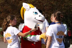 Dwie kobiety zwrócone tyłem do obiektywu. Między nimi „maskotka” biegu - koziołek. W tle zieleń drzew