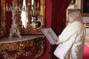 Od lewej: przy lustrze w zdobionej złotej ramie, na bogato zdobionej półce stoi porcelanowy kandelabr. Z prawej: przy umieszczonym na statywie wytłaczanym rysunku tego kandelabru stoi kobieta, która opuszkami palców dotyka wypukłości modelu