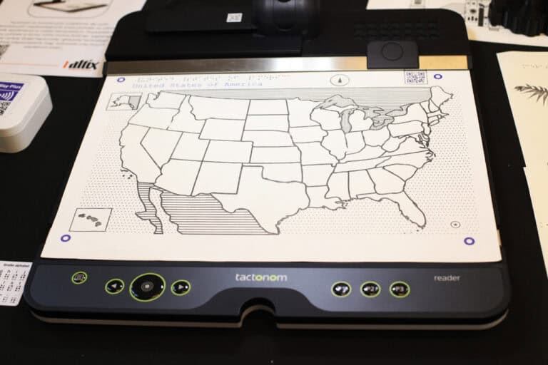 Zbliżenie na dotykową mapę Stanów Zjednoczonych, wydrukowaną czarnym konturem i liniami na białym tle, umieszczoną na urządzeniu zwanym "tactonom”. Nad mapą widnieje napis "United States of America" oraz tekst w alfabecie Braille’a