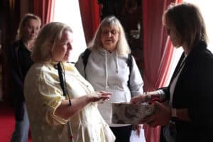 Na pierwszym planie dwie kobiety stojące naprzeciwko siebie. Kobieta z lewej dotyka palcami trzymany w dłoni mały przedmiot. Kobieta z prawej trzyma przed nią otwarte okrągłe pudełko. Scenie przygląda się kobieta będąca lekko w tle. Wszystkie stoją na tle okien z drapowanymi zasłonami w barwie łososiowej