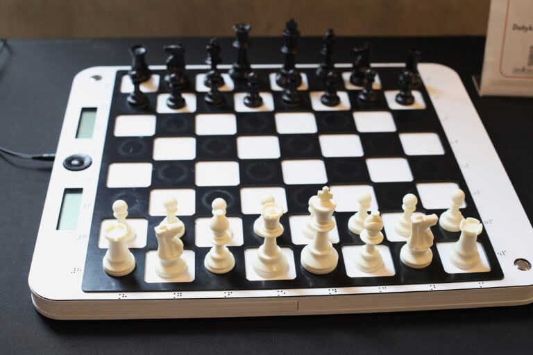 Zbliżenie na interaktywną szachownicę. Czarne i białe figury szachowe są ustawione w początkowych pozycjach, ale nie wszystkie figury stoją na odpowiednich miejscach. Po lewej stronie szachownicy widoczne są dwa małe ekrany cyfrowe i przyciski sterujące