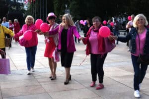 Plac Litewski w Lublinie. Zbliżenie na kilka kobiet, które w tańcu trzymają się za ręce. Każda z kobiet ma na sobie różowe elementy garderoby, niektóre trzymają różowe balony. W tle zieleń drzew i inni uczestnicy zgromadzenia