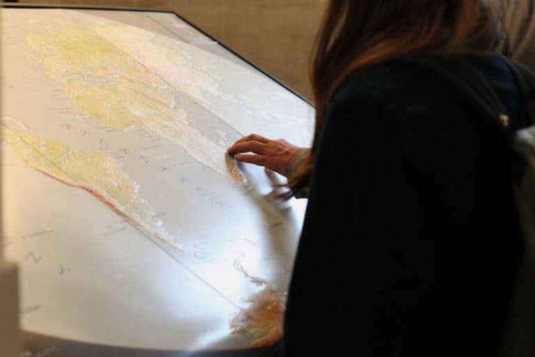 Osoba w ciemnej bluzie z kapturem pochyla się nad dużą interaktywną, dotykową mapą świata, na której widać dłoń dotykającą palcami powierzchni mapy w rejonie Afryki. Mapa ma wypukłe elementy i częściowo pada na nią światło