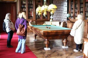 Wnętrze sali pałacowej. Na środku stoi wielki drewniany, prostokątny stół na grubych rzeźbionych nogach. Stół pokryty jest zielonym suknem. Nad nim wisi wielka zdobiona lampa z trzema półokrągłymi czaszami. Wokół stołu, z dwóch jego stron stoją cztery kobiety. W tle wielkie, drewniane regały, wypełnione książkami. Podłoga to drewniana mozaika