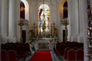 Wnętrze kaplicy. Półmrok. Na wprost wielki zdobiony witraż, przez który wpada światło. Widok na ołtarz główny, do którego prowadzi czerwona wykładzina. Po obu stronach wnętrza kaplicy rzędy krzeseł z miękkim, czerwonym obiciem. W pierwszych rzędach siedzą przodem do siebie cztery kobiety