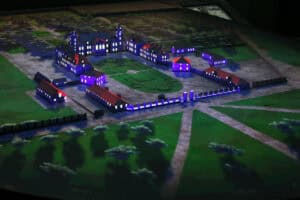 Półmrok. Widok z góry na podświetloną makietę zabudowań pałacowych i okolicy. Zabudowania są w barwach niebieskich, z podświetlonymi oknami i czerwono brązowymi dachami. Dookoła zieleń, poprzecinana liniami dróg i ciągów komunikacyjnych. Na pierwszym planie i w tle miniatury drzew