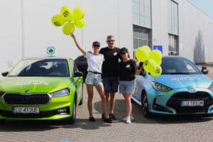 Pomiędzy dwoma samochodami trzyosobowa grupa z Uniwersytetu Jazdy: dwie kobiety i mężczyzna. Kobiety stoją po bokach mężczyzny i unoszą w górę żółte balony. W tle ściana budynku