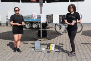 Punkt partnera zawodów: Głuche Miasto. Dwie kobiety ubrane na czarno. Pomiędzy nimi, na chodniku ekspozycja z ulotek i kart do nauki języka migowego