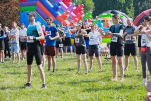 W kadrze zawodnicy biegu w strojach sportowych, z numerami startowymi przyczepionymi do koszulek, podczas rozgrzewki. W tle kolorowa, nadmuchiwana zjeżdżalnia dla dzieci