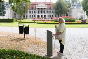 Na tle pałacu i ogrodu usytuowanego tuż przed nim, stoi kobieta trzymająca w prawej dłoni białą laskę. Jej lewa dłoń spoczywa na tarczy terminala z audioprzewodnikiem