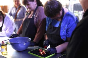 Kobieta w niebieskiej bluzce i czarnym fartuchu kuchennym pochyla się nad deską do krojenia. Na desce leżą cząstki jabłka. Obok deski stoi plastikowa, niebieska miska
