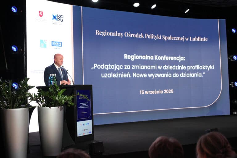 Widok na scenę. Od lewej, za mównicą mężczyzna (Jarosław Stawiarski) przy mikrofonie. Reszta kadru, to widok na ekran multimedialny z napisem: "Regionalny Ośrodek Polityki Społecznej w Lublinie. Regionalna Konferencja: 'Podążając za zmianami w dziedzinie profilaktyki uzależnień. Nowe wyzwania do działania.' 15 września 2025”