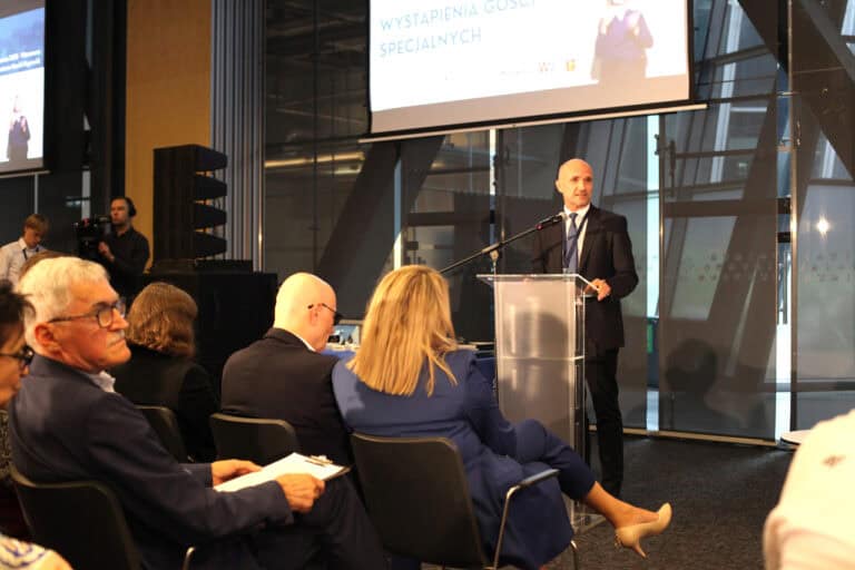 We wnętrzu sali konferencyjnej przy mównicy stoi mężczyzna w garniturze. Za nim znajduje się część ekranu projekcyjnego