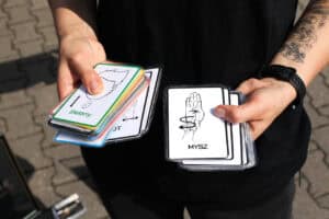 Zbliżenie na dłonie, a w nich karty do nauki języka migowego. Od lewej karta z napisem „zielony”, z prawej: „mysz”