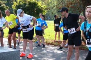 Na parkowej alei zawodnicy biegu w strojach sportowych, z numerami startowymi przyczepionymi do koszulek, podczas rozgrzewki. W tle zieleń roślin