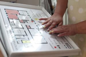 Dłonie spoczywają na legendzie wytłaczanego planu pomieszczeń. Wyraźne uwypuklenia i opisy w alfabecie Braille’a