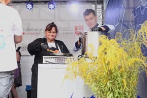 Za stanowiskiem do gotowania stoi od lewej: kobieta, która drewnianą łyżkę trzyma nad patelnią; obok mężczyzna w uniformie szefa kuchni, dłonią w czarnej rękawiczce trzyma pokrywkę dużego, metalowego garnka. W prawym dolnym rogu bukiet z żółtej nawłoci