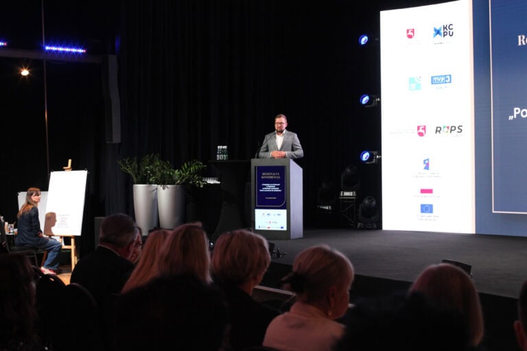 Wnętrze sali konferencyjnej, widok na scenę. Półmrok. Po lewej, przy mównicy mężczyzna w okularach podczas przemówienia. Z prawej fragment ekranu multimedialnego