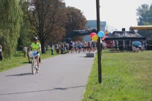Na pierwszym planie z lewej kobieta w kasku i odblaskowej kamizelce, jedzie na rowerze. W pewnej odległości za nią zawodnicy w strojach sportowych uchwyceni w biegu po przekroczeniu linii Startu. Otoczenie parku: drzewa z lewej, trawnik z prawej