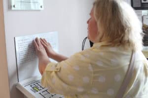 Kobieta dotyka palcami tablicy informacyjnej napisanej alfabetem Braille’a, która umieszczona jest na jasnej ścianie. Kobieta ma blond włosy i żółtą bluzkę w białe grochy
