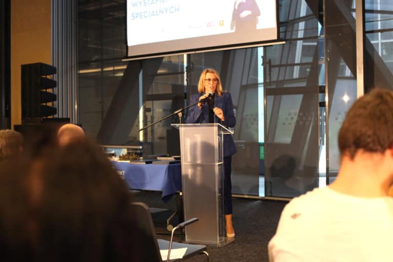 Kobieta w okularach przemawia na podium do siedzącej publiczności w sali konferencyjnej. Za nią znajduje się ekran projekcyjny z niewyraźną prezentacją
