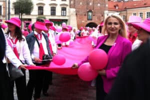 Plac Króla Władysława Łokietka. Kobieta w różowej marynarce i czarnej bluzce uśmiecha się, trzymając dwa różowe balony. Stoi na pierwszym planie zgromadzenia. Za nią duża grupa ludzi, głównie kobiet w różowych kapeluszach i różowych elementach stroju, trzyma razem szeroki pas różowego materiału. Tło stanowią zabytkowe kamienice i ceglana brama miejska (Brama Krakowska)