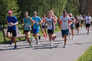 Zawodnicy w kolorowych strojach sportowych uchwyceni w biegu. Biegną po asfaltowej parkowej alei. W tle zieleń drzew