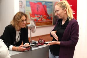 Dwie kobiety. Pierwsza od lewej opiera się o wysoki kontuar, trzyma w dłoni urządzenie (audioprzewodnik). Druga kobieta stoi po przeciwnej stronie kontuaru, w lewej ręce trzyma audioprzewodnik. W tle obraz z przewagą koloru czerwonego