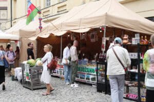 Stoisko Azerbejdżanu. Przy zadaszeniu powiewa flaga tego państwa. Wokół stoiska zgromadzeni ludzie i metalowy stół, na którym leżą arbuzy