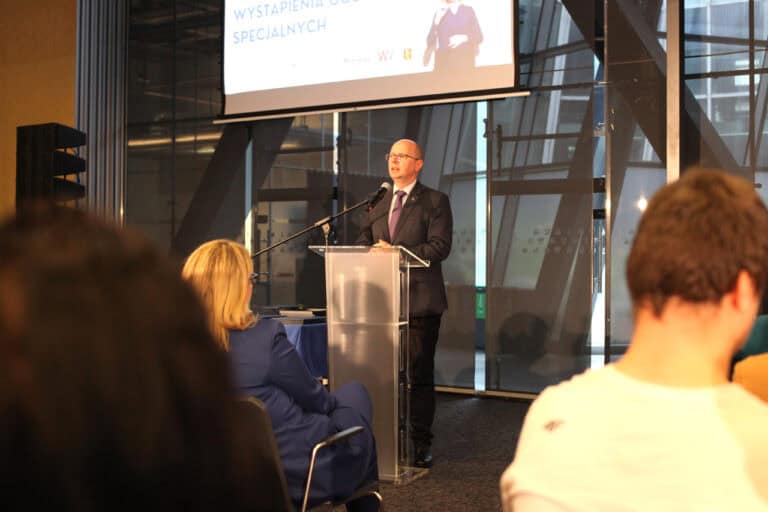 We wnętrzu sali konferencyjnej przy mównicy stoi mężczyzna w garniturze i w okularach. Za nim znajduje się część ekranu projekcyjnego. Przed mężczyzną widać nieostre sylwetki zgromadzonych gości