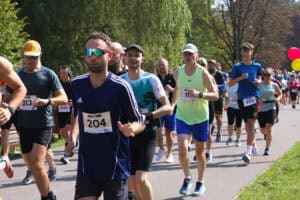 Z lewej zbliżenie na mężczyznę w przeciwsłonecznych okularach sportowych, z numerem 204. Za nim pozostali zawodnicy w kolorowych strojach sportowych uchwyceni w biegu