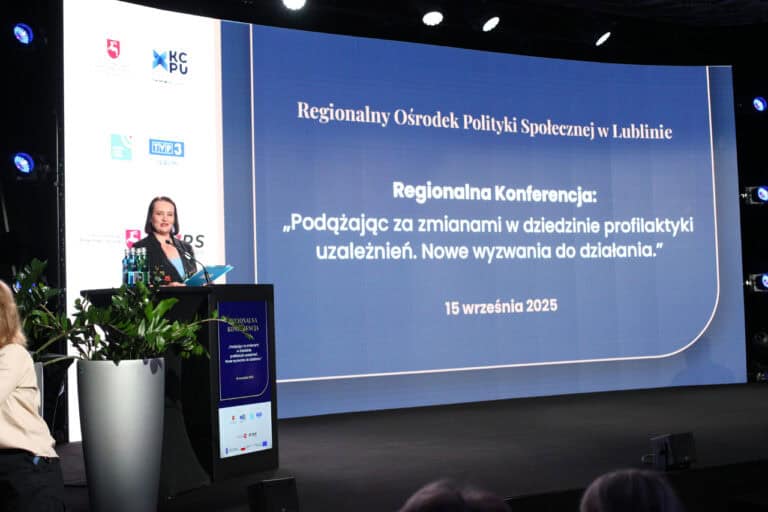 Kobieta przemawiająca na scenie; w tle duży ekran multimedialny, na którym wyświetlany jest napis: "Regionalny Ośrodek Polityki Społecznej w Lublinie. Regionalne Konferencja: 'Podążając za zmianami w dziedzinie profilaktyki uzależnień. Nowe wyzwania do działania.' 15 września 2025”