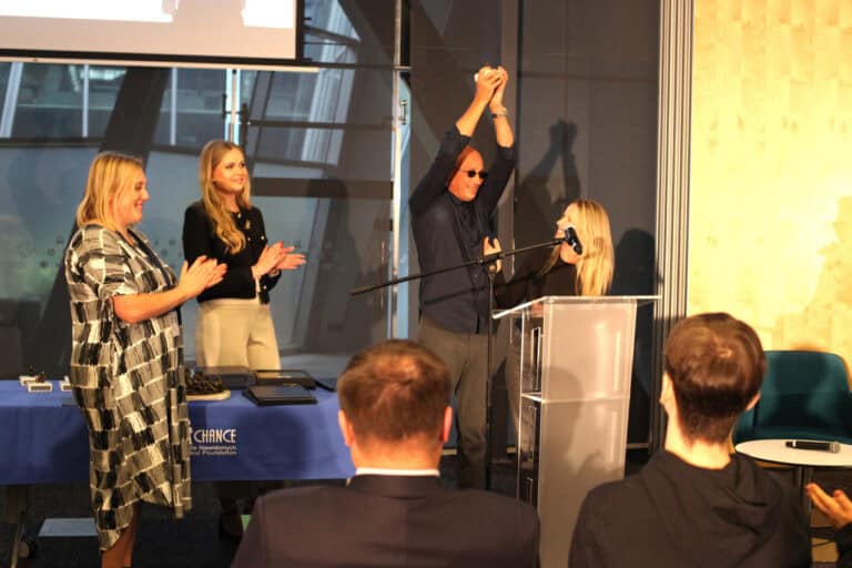 Cztery osoby na scenie podczas ceremonii. Mężczyzna w ciemnych okularach podnosi ręce w geście triumfu, stojąc za podium z mikrofonem, obok uśmiechniętej kobiety. Dwie inne kobiety stoją klaszcząc przy stole z niebieskim obrusem