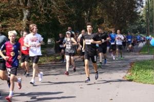 Zawodnicy w kolorowych strojach sportowych uchwyceni w biegu. Biegną po asfaltowej parkowej alei. W tle zieleń drzew