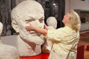 Od lewej okazała rzeźba (głowa) w jasnej barwie. Przed nią kobieta, która trzyma dłonie na rzeźbie. W tle wnętrze sali wystawienniczej