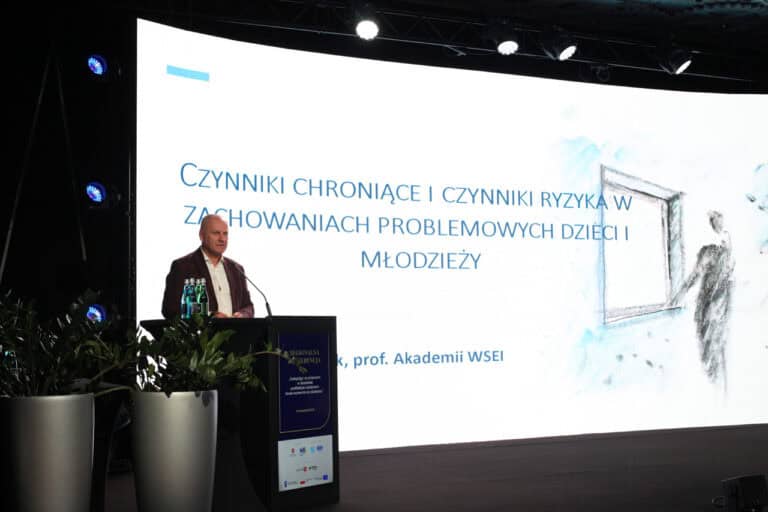 Mężczyzna (dr Wiesław Poleszak, prof. WSEI) przemawiający na scenie; w tle duży ekran, na którym wyświetlany jest napis: „Czynniki chroniące i czynniki ryzyka w zachowaniach problemowych dzieci i młodzieży”