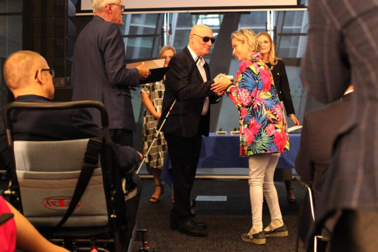 Na scenie odbywa się ceremonia. Mężczyzna w garniturze i ciemnych okularach, trzymający białą laskę (Marek Kalbarczyk), wręcza mały przedmiot kobiecie w kolorowej kwiecistej marynarce. Z tyłu widoczne są dwie inne osoby i mężczyzna trzymający dyplom. Na pierwszym planie, z lewej strony, widoczna jest osoba siedząca na wózku inwalidzkim