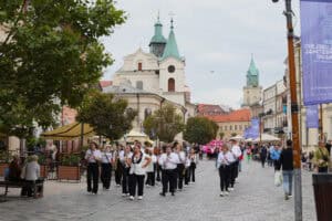 Orkiestra dęta maszeruje po brukowanym Deptaku w Lublinie, prowadząc paradę. Uczestnicy orkiestry są ubrani w białe koszule i czarne spodnie/spódnice, niektórzy niosą instrumenty dęte. W tle widoczna jest historyczna architektura. Po lewej stronie widać ławki i drzewa, a po prawej bilbordy reklamowe. Inni piesi znajdują się na dalszym planie