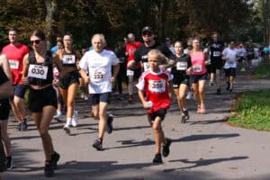 Na tle ciemnej zieleni drzew zawodnicy w kolorowych strojach sportowych uchwyceni w biegu. Biegną po asfaltowej parkowej alei