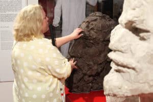 Od lewej kobieta, której dłonie spoczywają na ciemnej, okazałej rzeźbie w barwach brązu. Od prawej fragment innej rzeźby rozmyty w kadrze