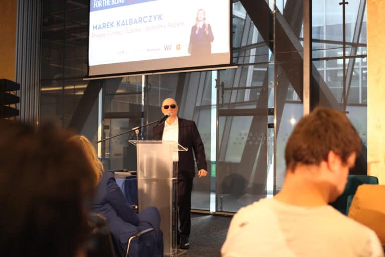 We wnętrzu sali konferencyjnej przy mównicy stoi mężczyzna w garniturze i ciemnych okularach. Za nim znajduje się część ekranu projekcyjnego, na którym widoczny jest napis: „Marek Kalbarczyk. Prezes Fundacji Szansa - Jesteśmy Razem”