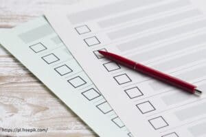 Czerwony długopis spoczywa na dwóch kartkach z listami kontrolnymi i polami wyboru, które leżą na jasnej drewnianej powierzchni.