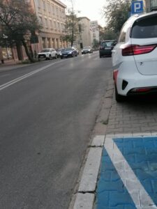 Miejsce parkingowe dla osób z niepełnosprawnościami przy skrzyżowaniu ulic Chopina i Krakowskiego Przedmieścia; widok na ulice Chopina. Fragment chodnika z wymalowanym biało-niebieskim oznaczeniem miejsca parkingowego. Przed nim parkuje białe auto; w tle znak drogowy "P" oraz jezdnia i zabudowania