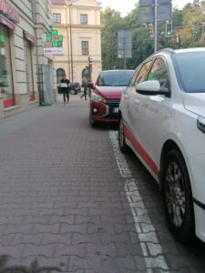 Miejsce parkingowe dla osób z niepełnosprawnościami przy skrzyżowaniu ulic Chopina i Krakowskiego Przedmieścia. Widok na Krakowskie Przedmieście. Ujęcie chodnika z perspektywy białego samochodu, za którym stoi krzywo zaparkowany pojazd osobowy, nie mieszczący się w wymalowanych liniach; w tle zabudowania.
