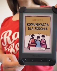 Kobieta trzyma czytnik e-booków, na którym widnieje okładka poradnika zatytułowany „Komunikacja dla zdrowia” z ilustracją przedstawiającą trzy stylizowane osoby rozmawiające.