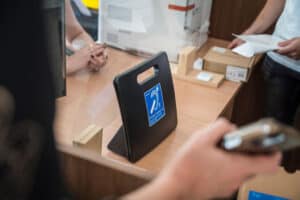 W kadrze ręka trzymająca telefon, a na stole znajduje się pętla indukcyjna z piktogramem przekreślonego ucha. W tle widać inne osoby i drukarkę.