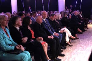 Grupa dorosłych osób w formalnych strojach siedzi w rzędach, obserwując wydarzenie poza kadrem. W pierwszym rzędzie widoczni są, m.in., kobieta w turkusowym żakiecie, mężczyzna w garniturze i krawacie, kobieta w czarnej i czerwonej sukience, mężczyzna w szacie duchownej oraz inni goście